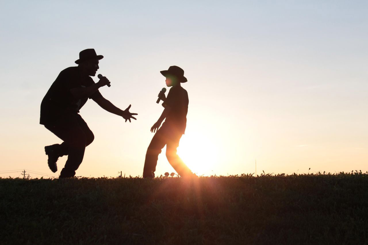 Ein junger und ein erwachsener Mann singen und tanzen in der Abendsonne.