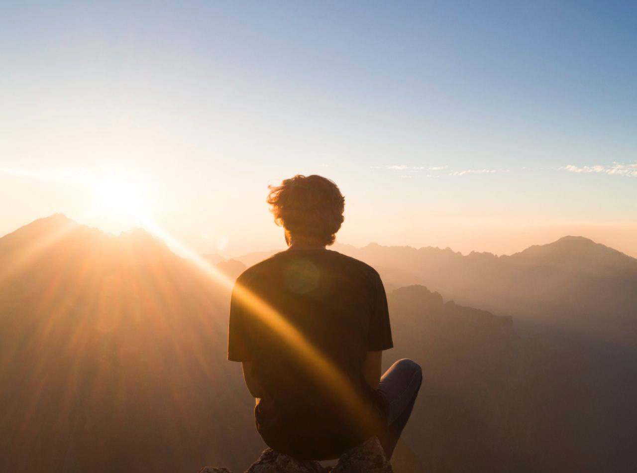 Ein Mann sitzt auf einem Gipfel und blickt in den Sonnenuntergang.